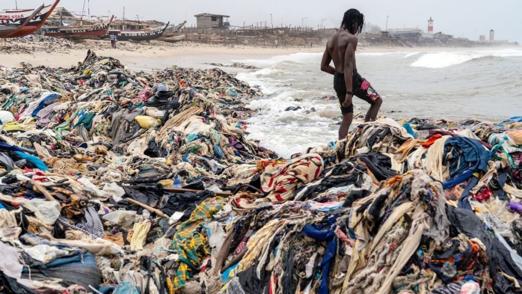 A man stands amidst piles of discarded clothing washed ashore on a beach in Ghana, highlighting the environmental impact of fast fashion. Photo by Muntaka Chasant.