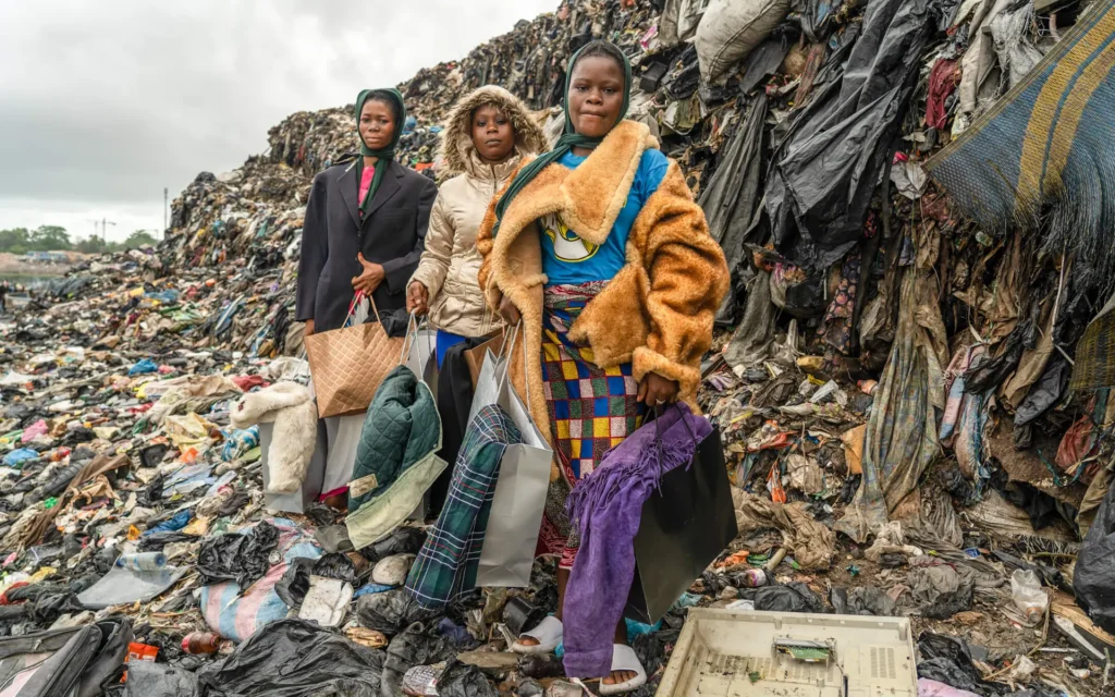 Three women stand in front of a towering pile of discarded clothing waste near Kantamanto Market in Ghana, a result of fast fashion pollution. Photo by The Revival.