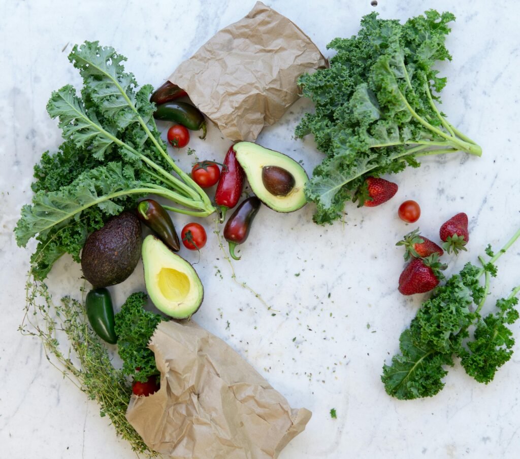 Fresh kale, avocado, strawberries, and other produce on a marble surface with paper bags.