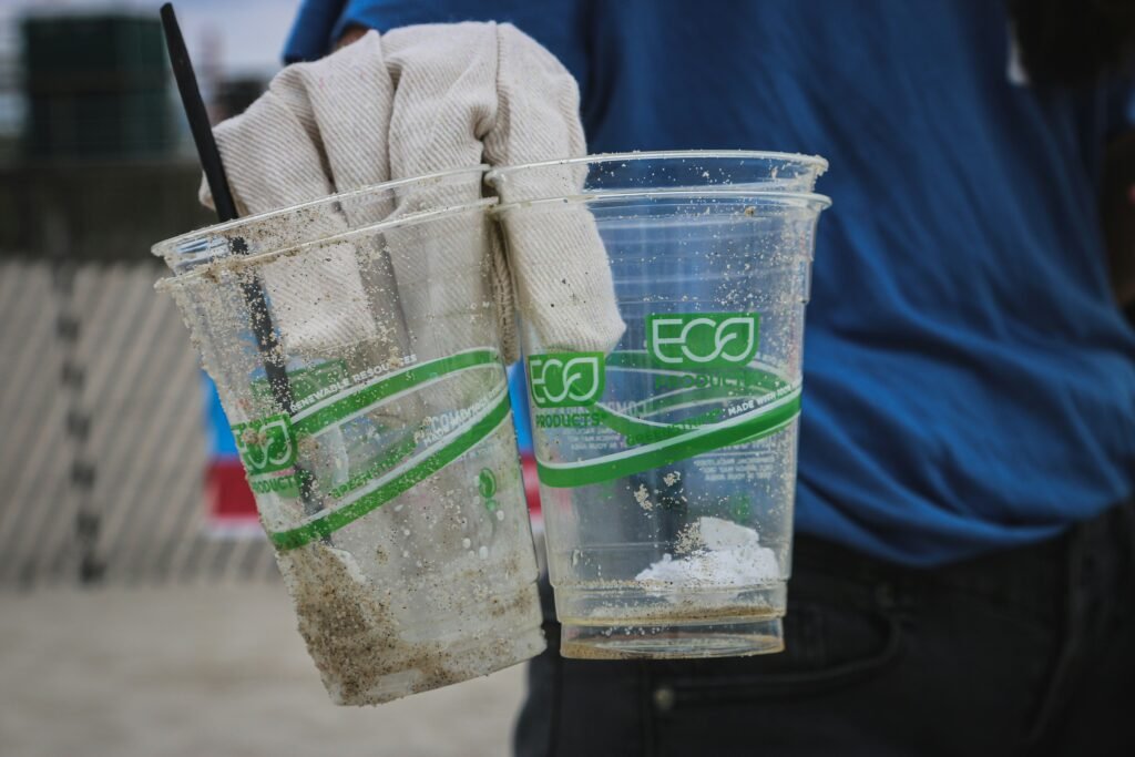 Two 'eco-friendly' plastic cups with green labels held by a gloved hand during a beach cleanup, exposing greenwashing.