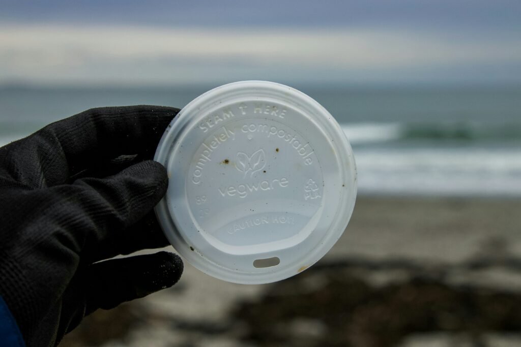 Compostable Vegware lid held by a gloved hand near the ocean, highlighting greenwashing in single-use plastics.